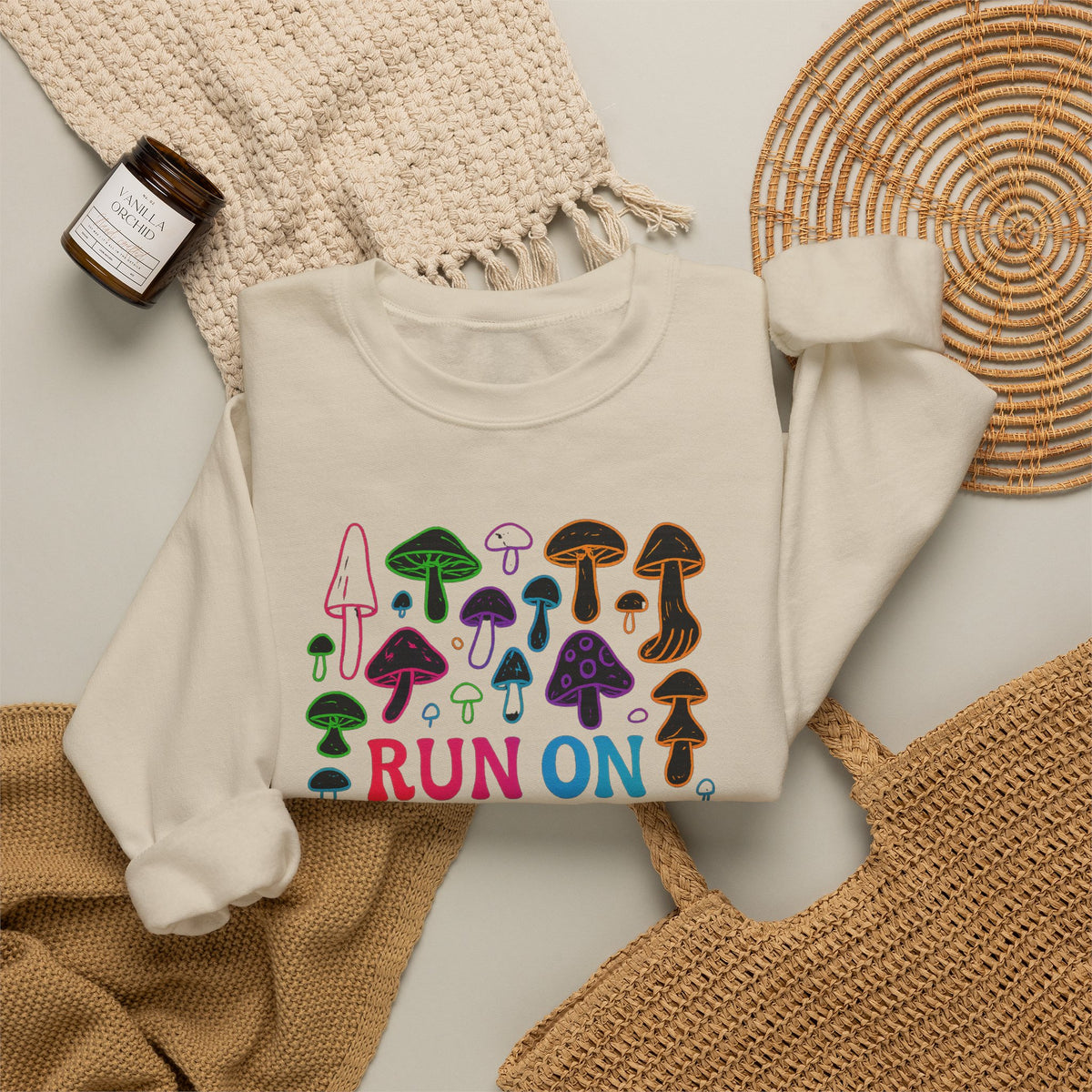 An off-white crewneck sweatshirt with a colorful mushroom graphic and the text 'RUN ON' printed on the front, styled in a flat lay with a woven blanket, a small candle, and a wicker tray.