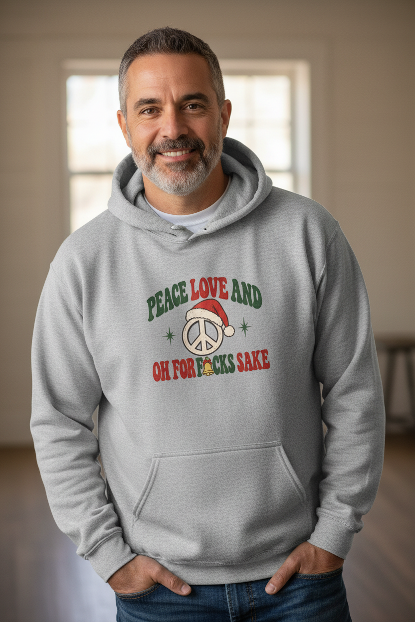A smiling man wearing a light gray hoodie with a festive graphic that reads 'PEACE LOVE AND OH FOR F*CKS SAKE' in green and red text around a peace sign with a Santa hat, standing in a softly lit indoor room.