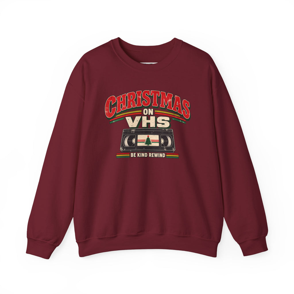 A maroon crewneck sweatshirt featuring a retro Christmas graphic on the chest that reads 'CHRISTMAS ON VHS' in red and cream lettering above a cassette tape illustration, with 'BE KIND REWIND' underneath.