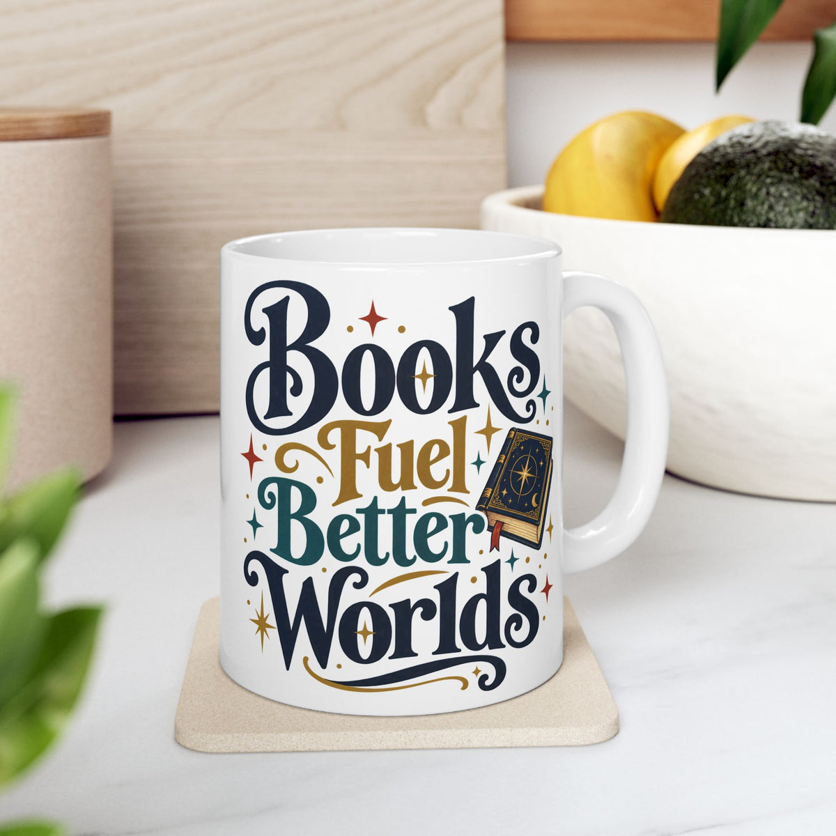 A white ceramic coffee mug with the phrase 'Books Fuel Better Worlds' in stylized navy, gold, and teal lettering, resting on a beige coaster on a kitchen counter.