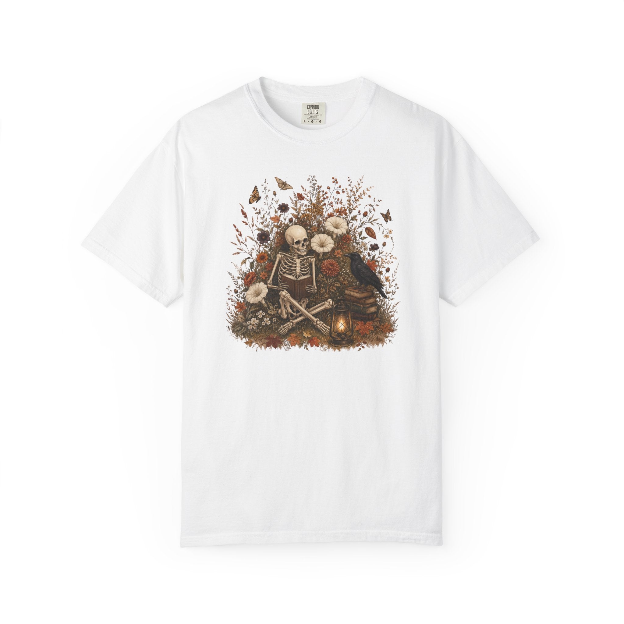 A white short-sleeve crewneck t-shirt featuring a detailed graphic print on the front. The graphic depicts a skeleton sitting on the ground surrounded by skulls, flowers, butterflies, and a raven, with a small glowing lantern at the bottom
