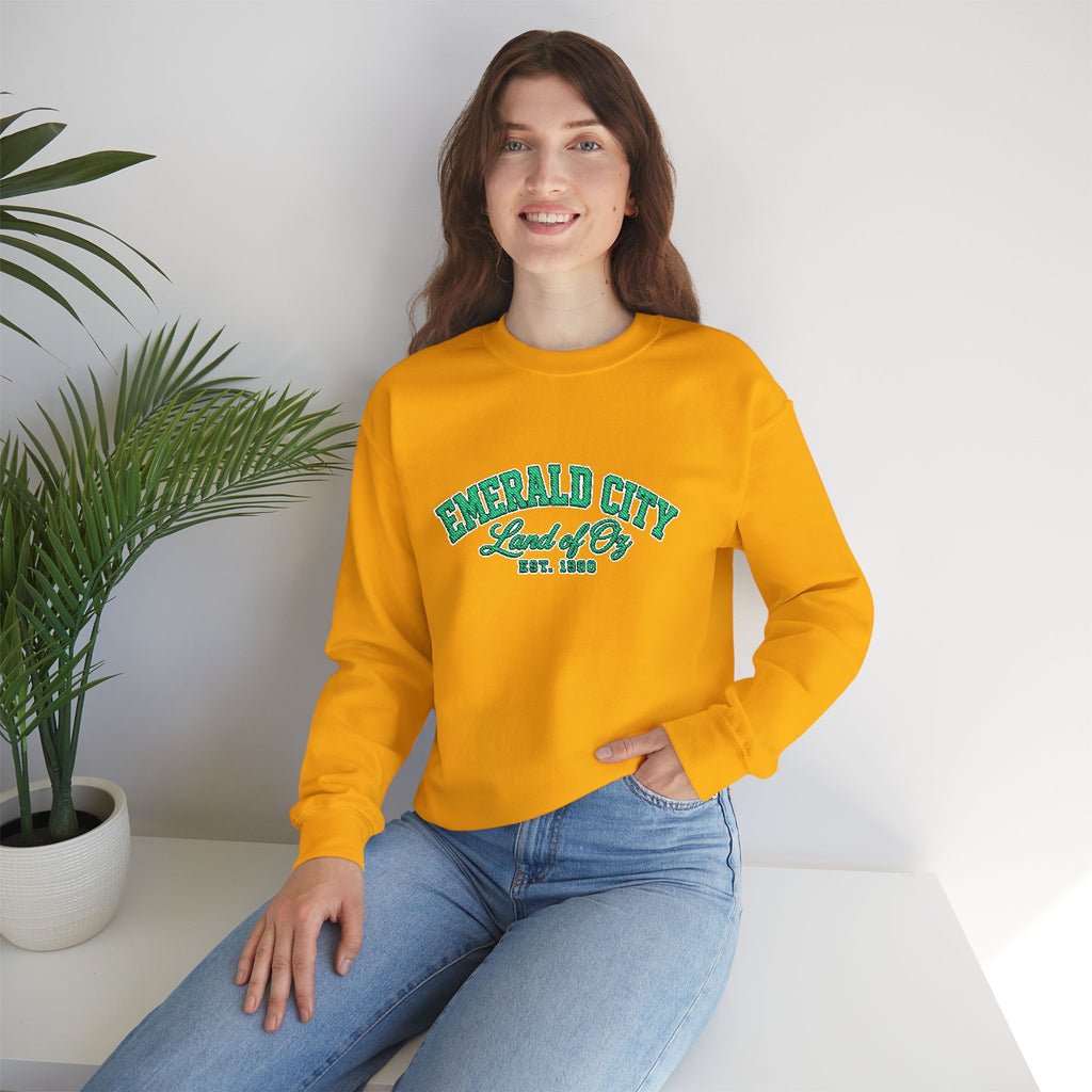 A woman wearing a bright yellow sweatshirt with 'EMERALD CITY Land of Op EST. 1900' printed in teal on the chest, paired with light blue jeans, sitting in a bright room next to a potted plant.