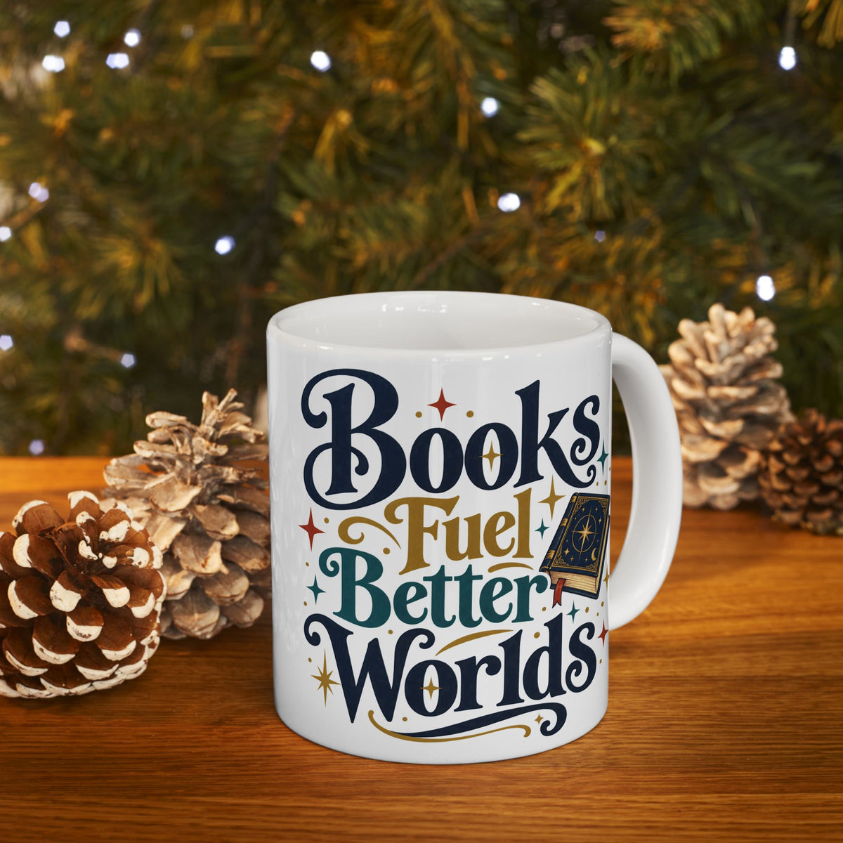 A white ceramic mug with the phrase 'Books Fuel Better Worlds' in stylized navy, gold, and teal lettering, placed on a wooden surface with pinecones and a blurred Christmas tree in the background.