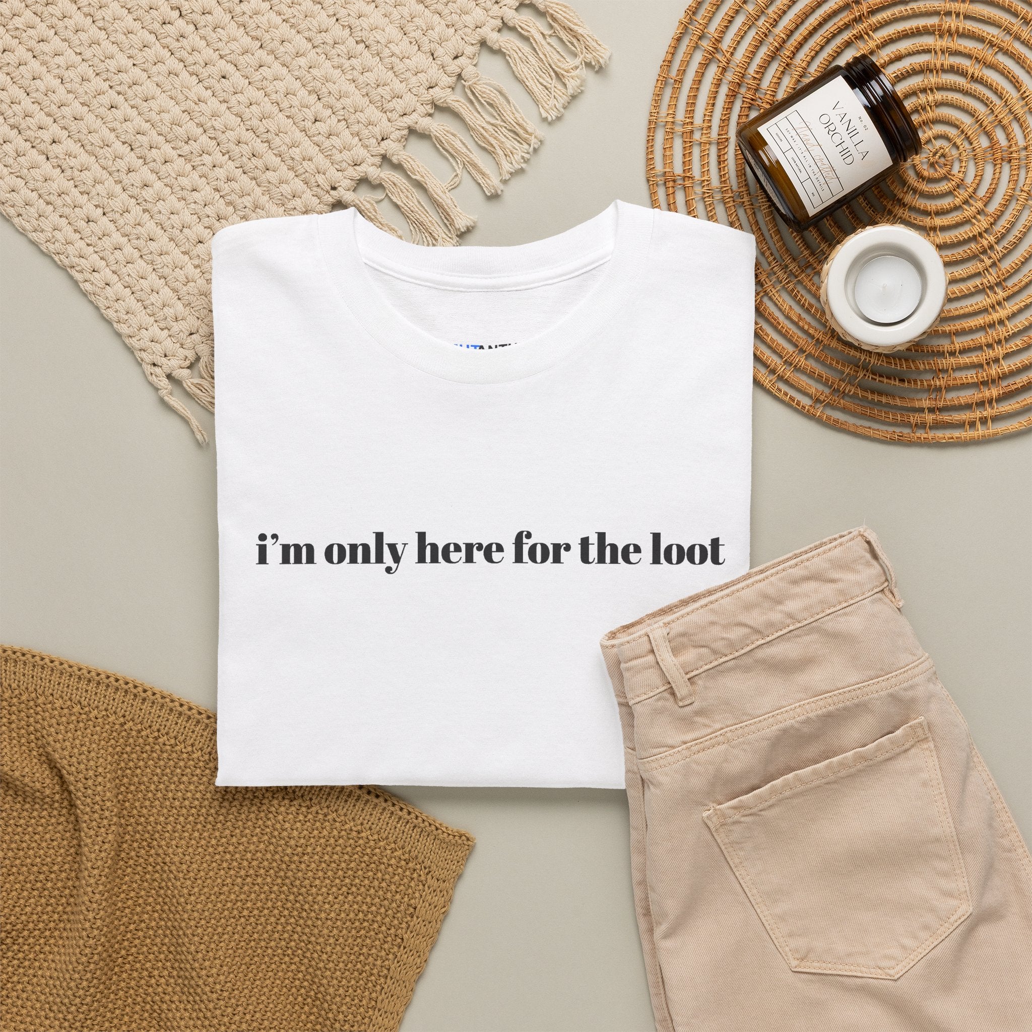 I'm Only Here for the Loot minimal text graphic tee in white, folded flat lay styled with a woven placemat and tan trousers