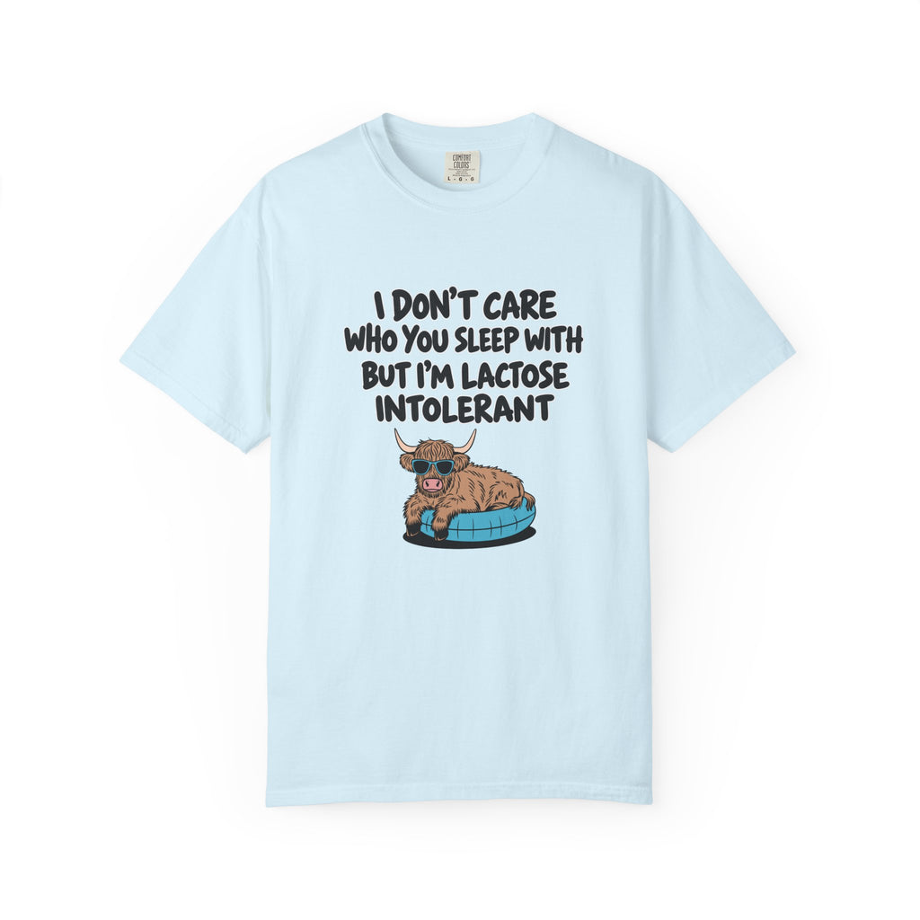 A light blue short-sleeve t-shirt with a humorous graphic on the front. The graphic features the text 'I DON'T CARE WHO YOU SLEEP WITH BUT I'M LACTOSE INTOLERANT' in black lettering above an illustration of a brown highland cow lying on a blue pet bed.