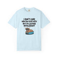 A light blue short-sleeve t-shirt with a humorous graphic on the front. The graphic features the text 'I DON'T CARE WHO YOU SLEEP WITH BUT I'M LACTOSE INTOLERANT' in black lettering above an illustration of a brown highland cow lying on a blue pet bed.