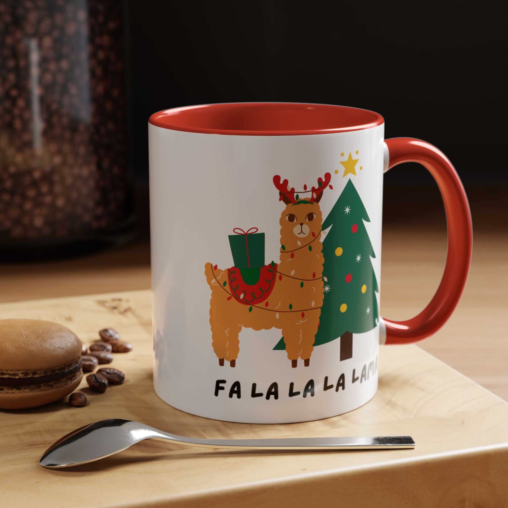 A white ceramic mug with a red interior and handle, featuring a festive holiday illustration of a llama wearing a reindeer helmet next to a Christmas tree, with the text 'FA LA LA LA LA' printed below. The mug is styled on a wooden surface with a spoon, coffee beans, and a macaron in the background.