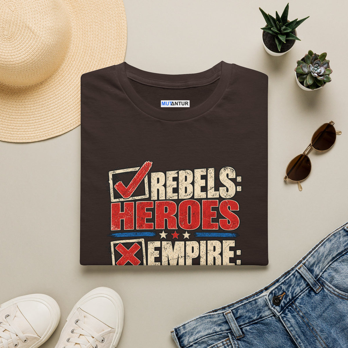 A folded dark brown graphic T-shirt is styled in a flat lay with a straw hat, white sneakers, blue jeans, sunglasses, and small potted succulents on a light beige background. The shirt features a distressed graphic that reads 'REBELS: HEROES' in red and white with a checkmark, and 'EMPIRE:' in red with an X.