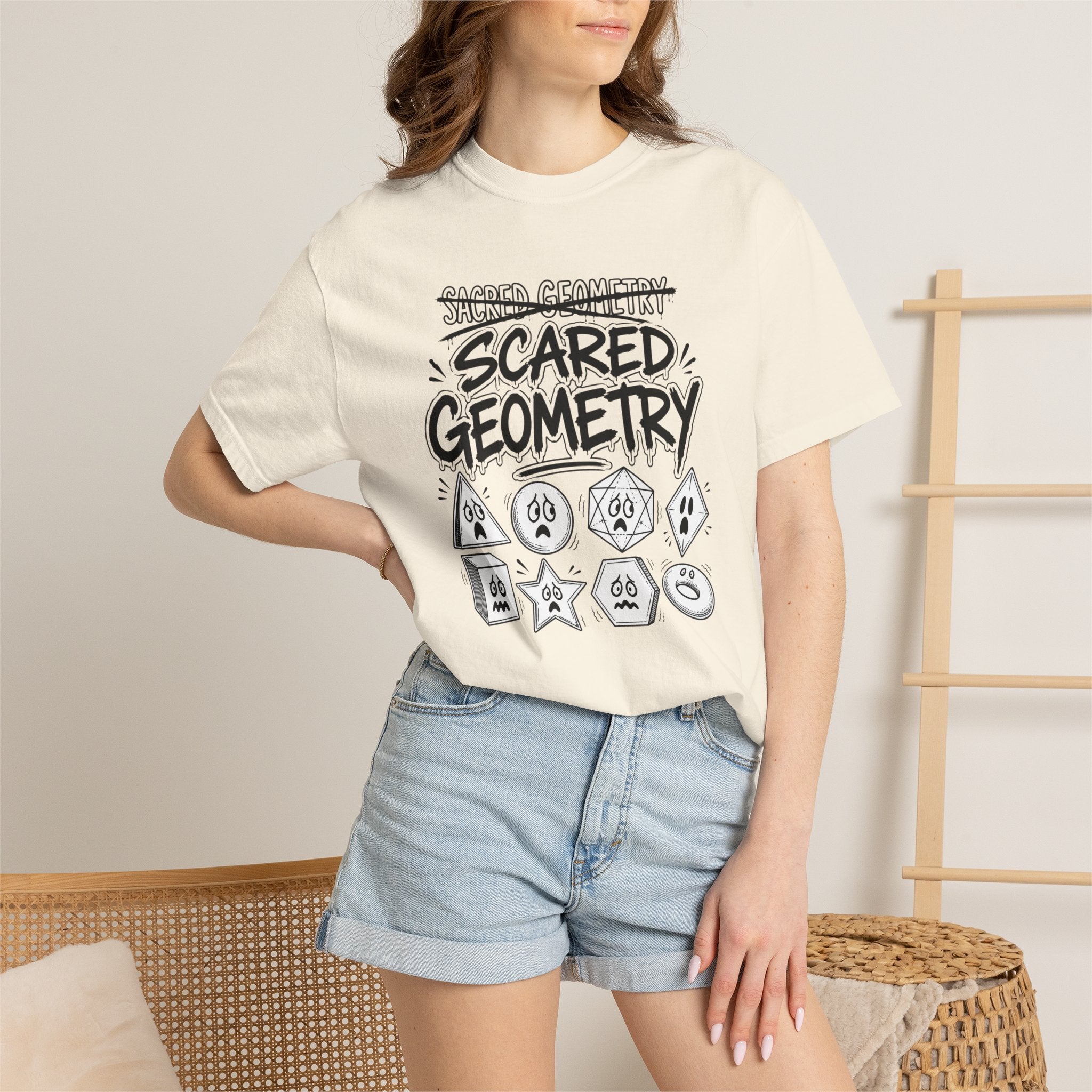 A woman wearing a butter-yellow 'Scared Geometry' graphic t-shirt with spooky-faced dice designs, paired with denim shorts, standing in front of a wicker chair.