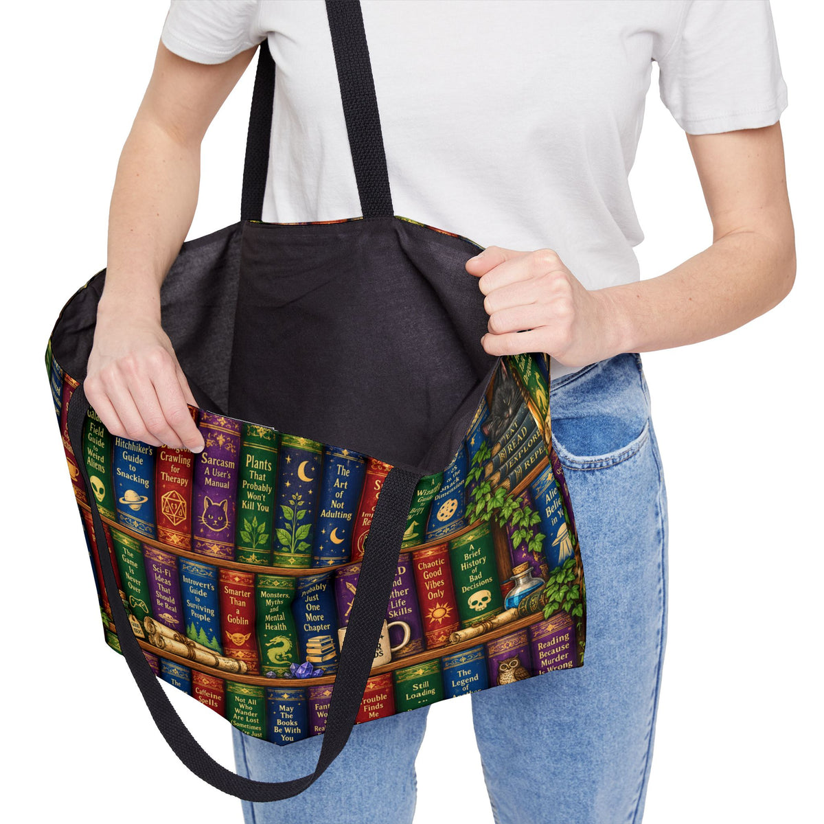A person wearing a white t-shirt and blue jeans holds up a large tote bag featuring a colorful printed design of fantasy book spines on a library shelf, with a black interior and black shoulder straps.