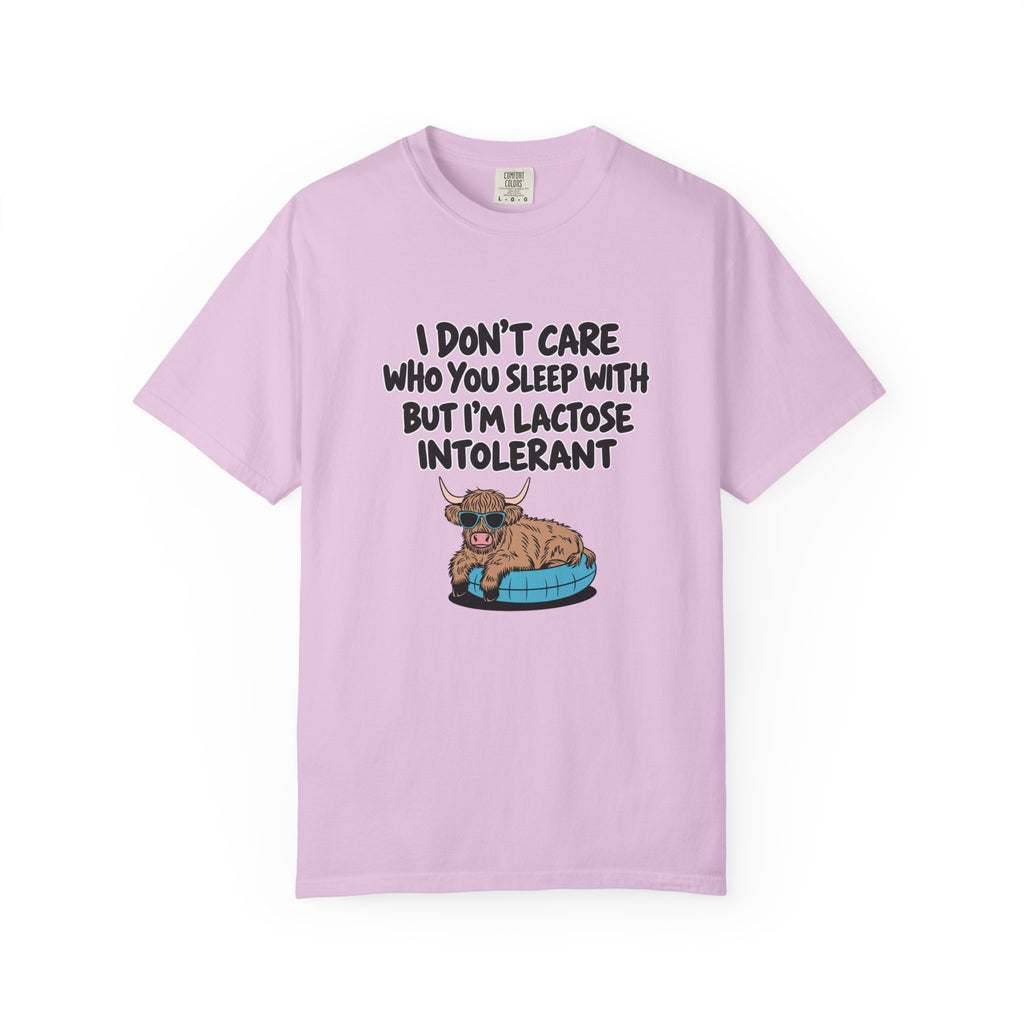 A light purple short-sleeve t-shirt with a humorous graphic on the front. The graphic features the text 'I DON'T CARE WHO YOU SLEEP WITH BUT I'M LACTOSE INTOLERANT' in black capital letters above an illustration of a brown highland cow lying on a blue and black tire.