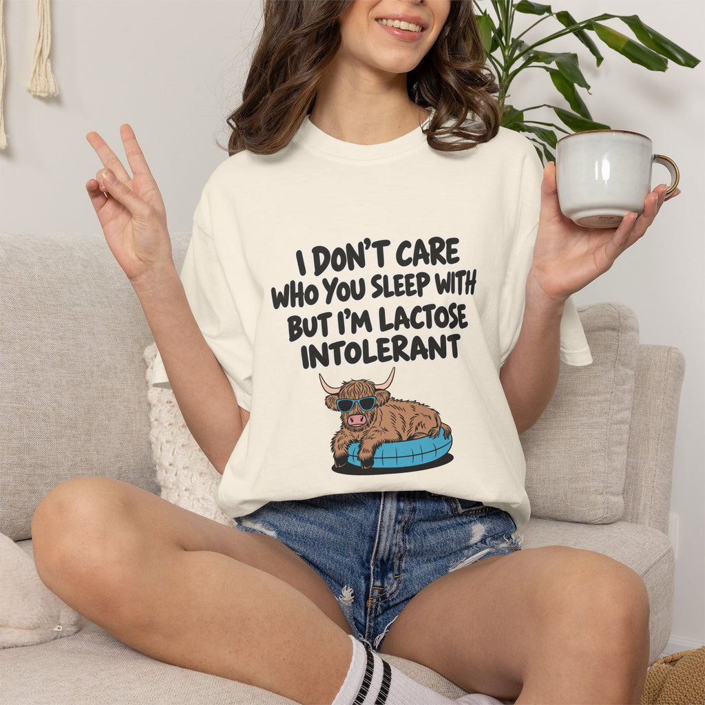A woman sitting on a light beige couch wearing a cream-colored graphic t-shirt with a cartoon cat and the text 'I DON'T CARE WHO YOU SLEEP WITH BUT I'M LACTOSE INTOLERANT'. She is holding a white mug in one hand and making a peace sign with the other, wearing denim shorts and white socks.