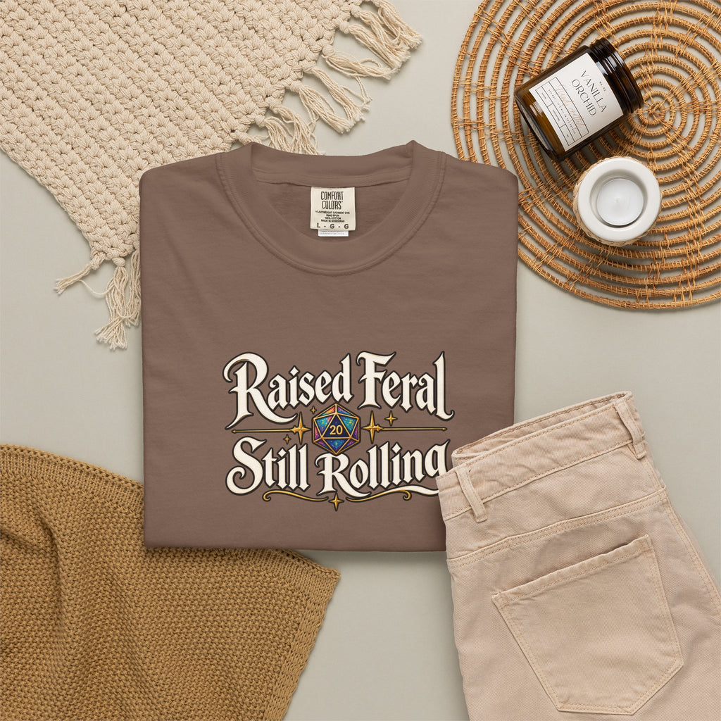 A folded taupe-brown graphic T-shirt is styled in a flat lay with beige pants, a woven blanket, a candle jar, and a small cup on a light neutral background. The shirt features the text 'Raised Feral Still Rolling' with a colorful 20-sided die graphic in the center.