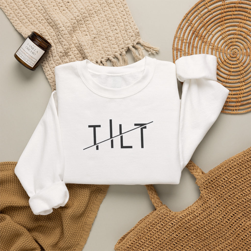 A folded white crewneck sweatshirt with  TILT' printed in black across the chest, styled in a flat lay with a woven blanket, a small candle jar, and a woven tray on a neutral background.