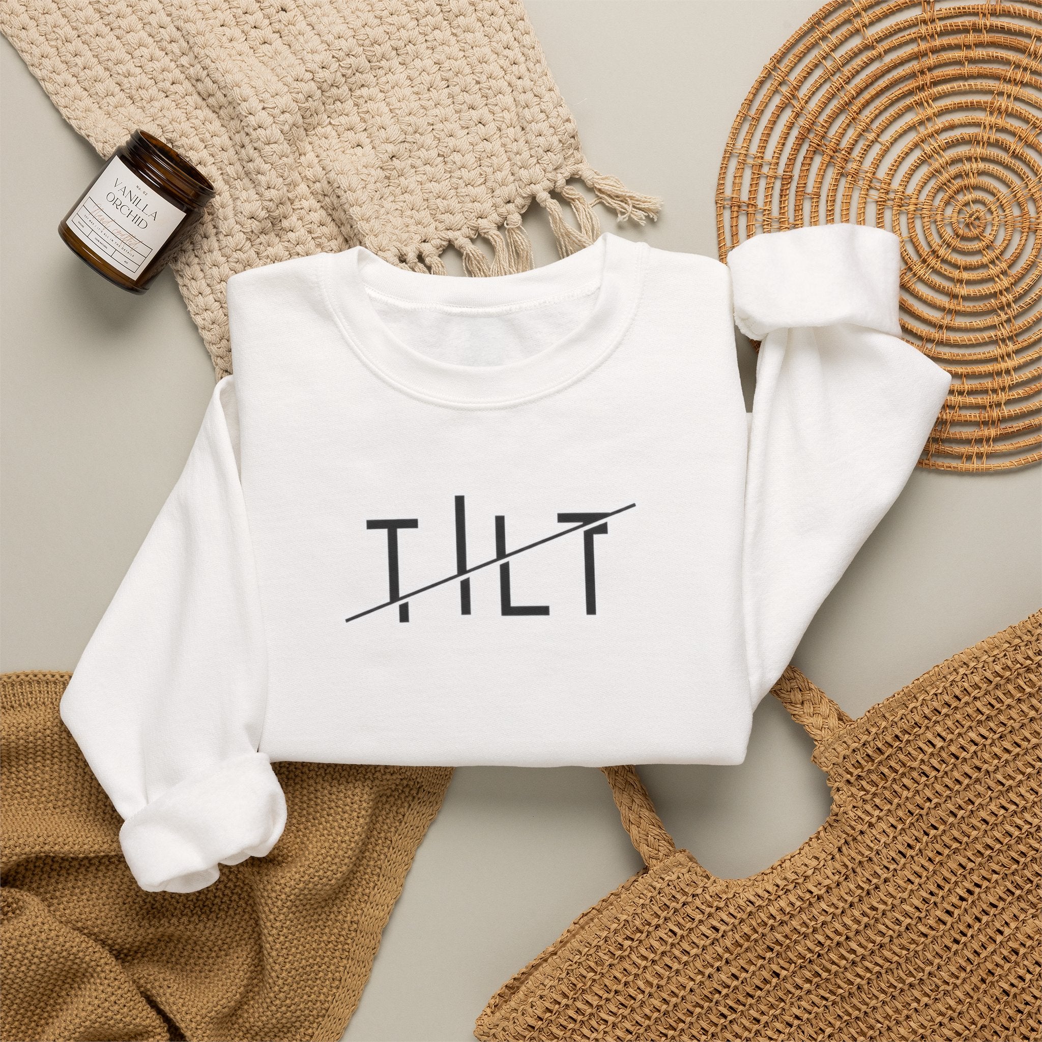 A folded white crewneck sweatshirt with  TILT' printed in black across the chest, styled in a flat lay with a woven blanket, a small candle jar, and a woven tray on a neutral background.