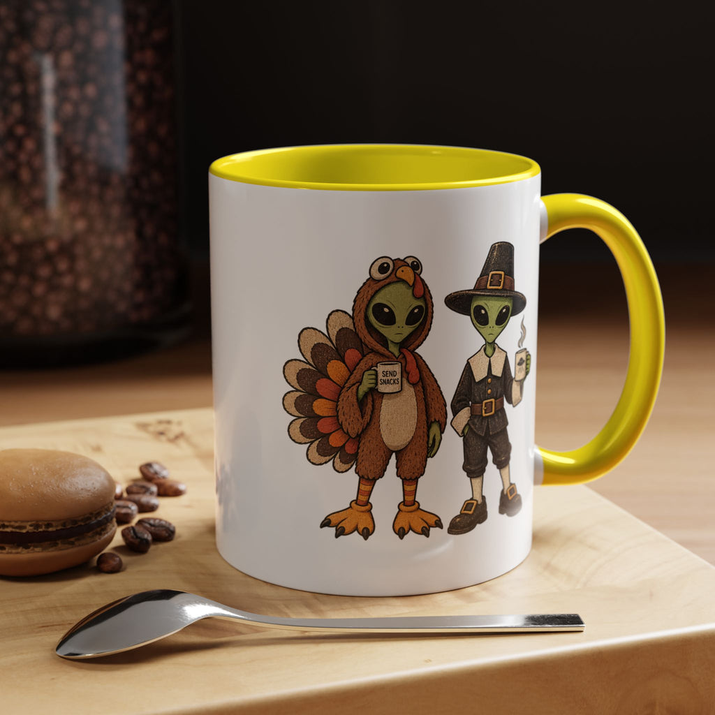 A white ceramic mug with a bright yellow interior and handle, featuring a cartoon illustration of a turkey and a pilgrim holding coffee cups. The mug is set on a wooden surface with a spoon in front and a blurred background.