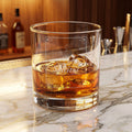 A close-up of a clear glass tumbler with amber liquid inside, featuring a frosted white engraved label that reads 'EX LIBRIS Spiritus' with an open book icon and decorative corner flourishes. This is placed on a marble bar counter
