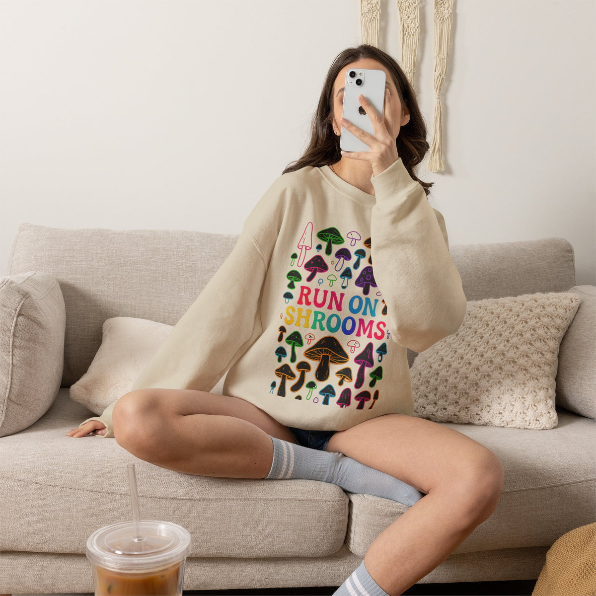 A woman takes a mirror selfie while sitting on a beige couch, wearing a cream sweatshirt with a colorful mushroom graphic and the text 'RUN ON SHROOMS'.