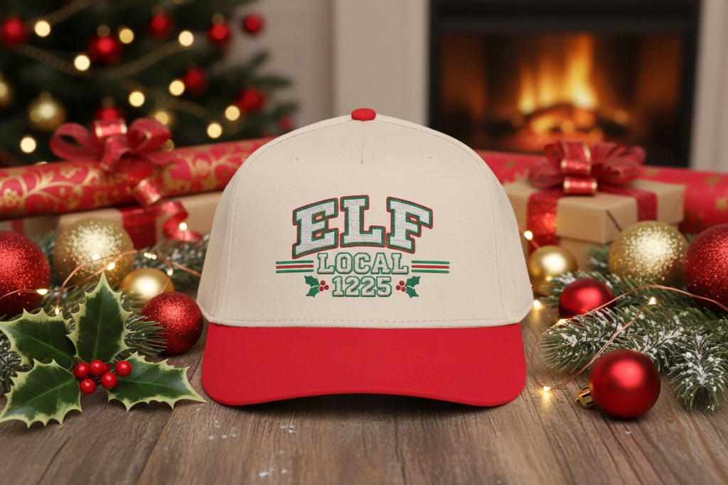 A red and off-white baseball cap with 'ELF LOCAL 1225' embroidered on the front, styled on a wooden table with Christmas decorations, wrapped gifts, and a warm fireplace in the background