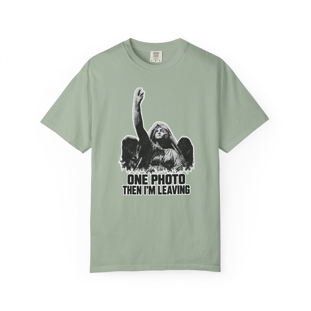 A sage green short-sleeve t-shirt with a black-and-white graphic of a woman raising her fist, accompanied by the text 'ONE PHOTO THEN I'M LEAVING'.