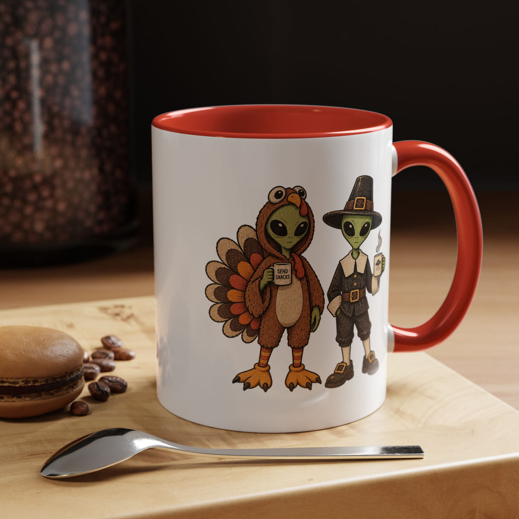 A white ceramic coffee mug with a red interior and handle, featuring a cartoon illustration of a turkey and a Pilgrim alien. The mug is set on a wooden surface next to a spoon and a macaron, with a blurred background showing coffee beans.