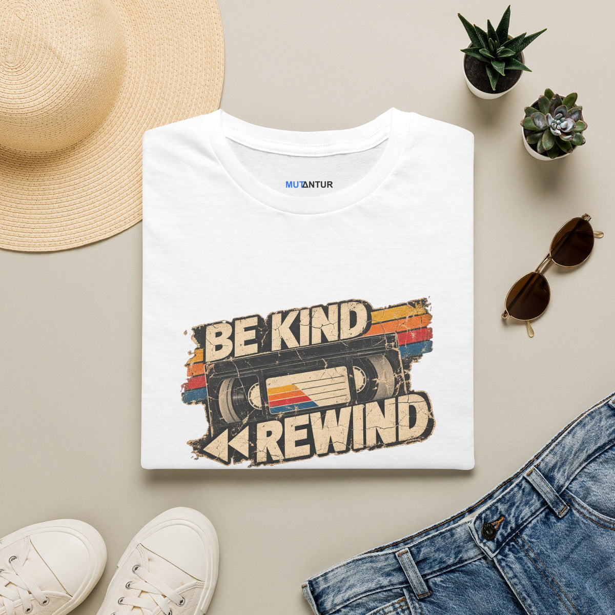 A folded white graphic T-shirt is styled in a flat lay with a straw hat, white sneakers, blue jeans, sunglasses, and small potted succulents on a light beige background. The shirt features a retro cassette-tape graphic with the words 'BE KIND REWIND' and a small visible neck print reading 'MUTANTUR'