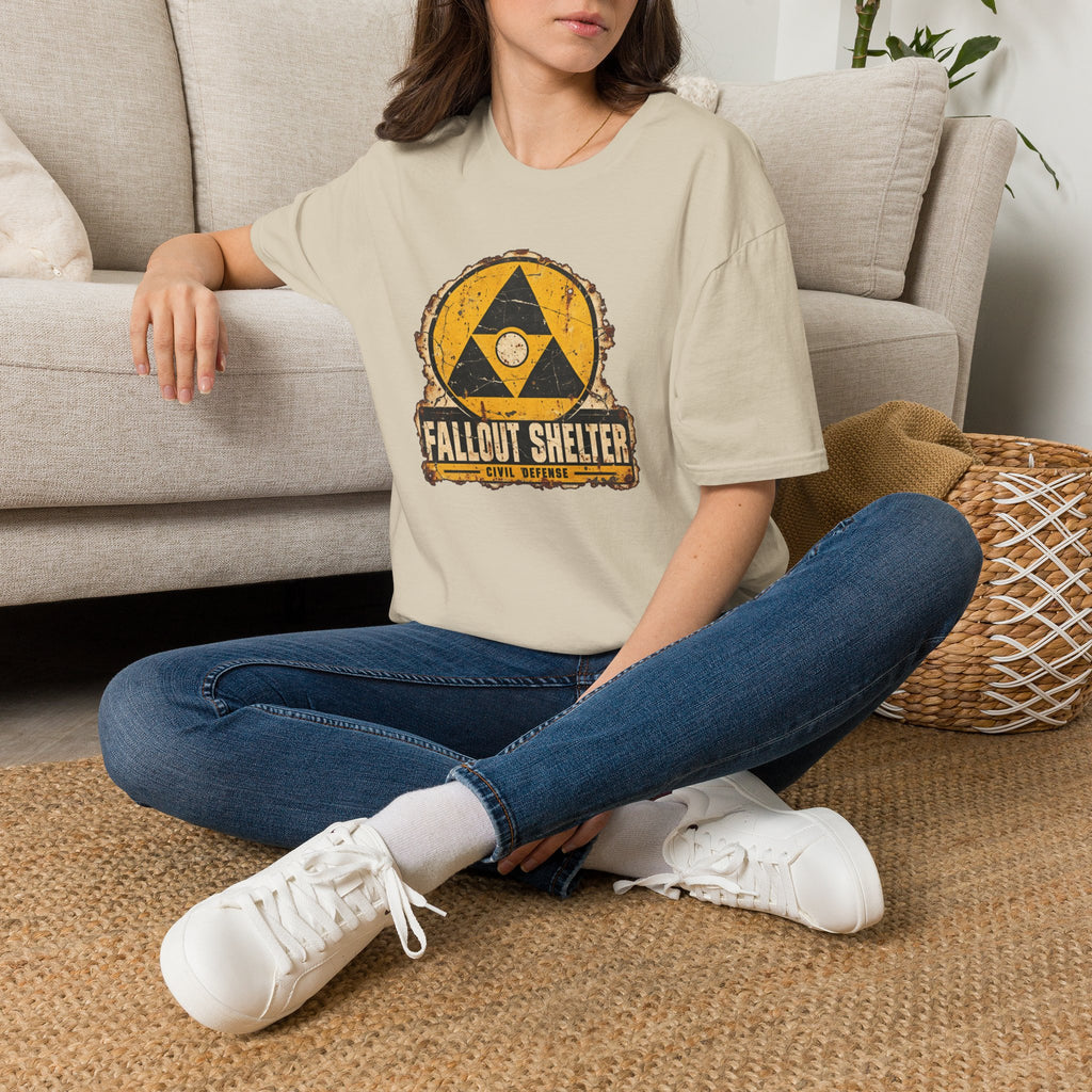 A sitting woman wearing a beige crew neck shirt with a vintage graphic depicting a  yellow and black  Fallout Shelter design with the words Civil Defense at the bottom